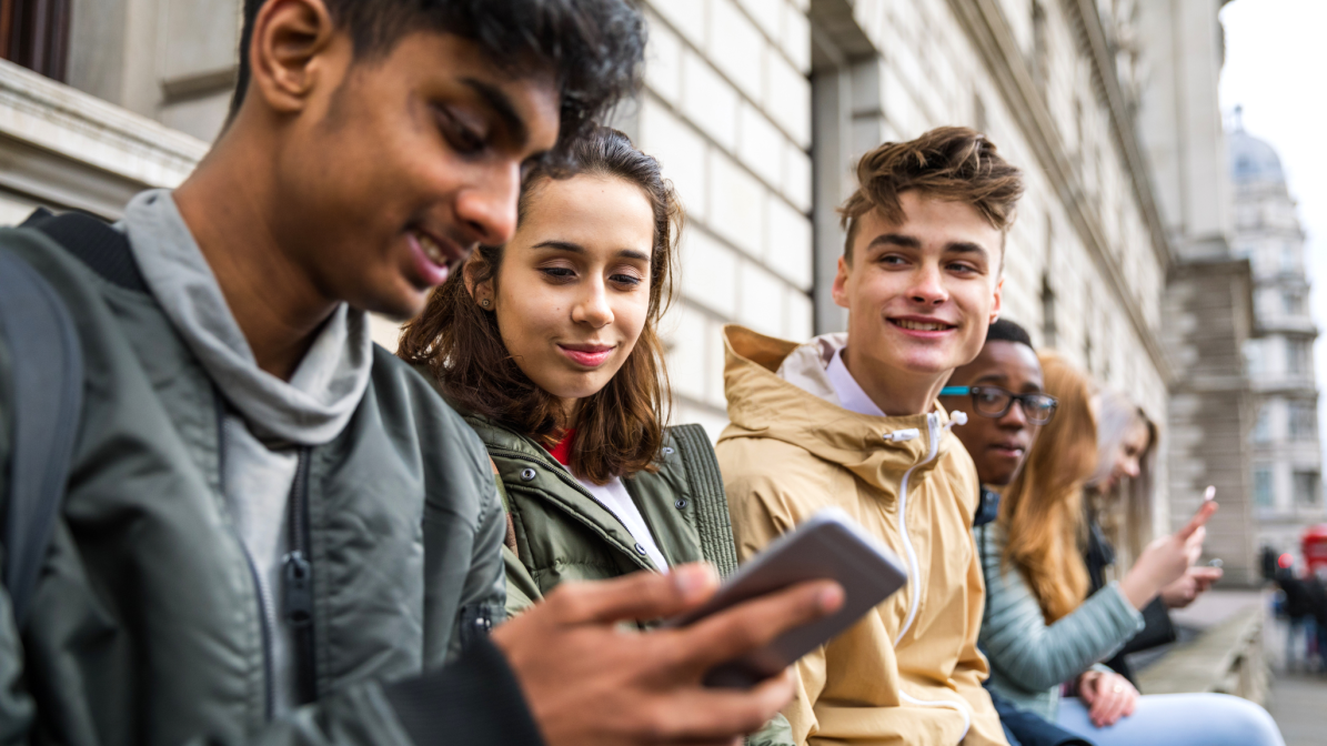 Brands_Image_1_Oct_25 Five students socialising on a city bench, one using a smartphone — a real-world glimpse into how next-gen consumers connect and engage, offering brands insight into reaching student audiences through mobile and social platforms.